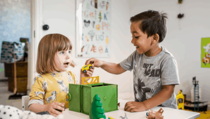 Two young children play with toy cars and animals at a table in a brightly decorated room, smiling and interacting with each other.